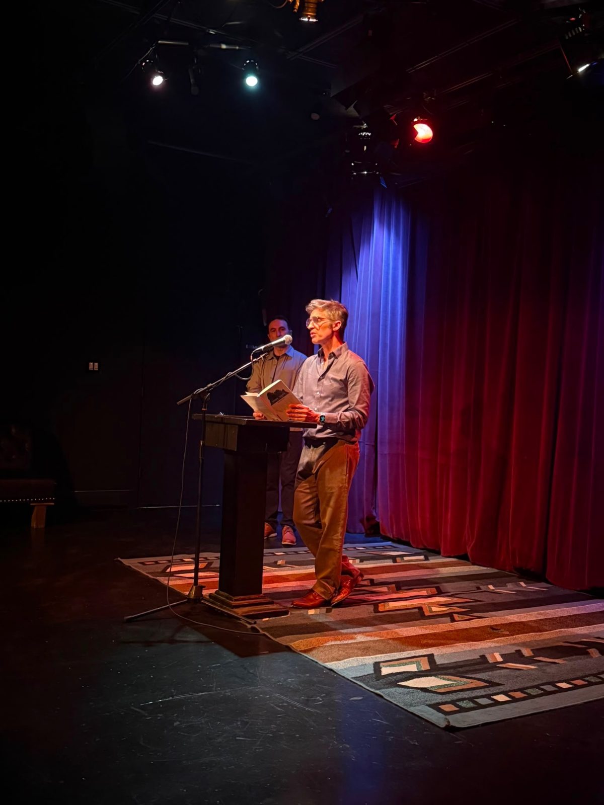 Blas Falconer reading from a book at the podium, a fellow reader behind him
