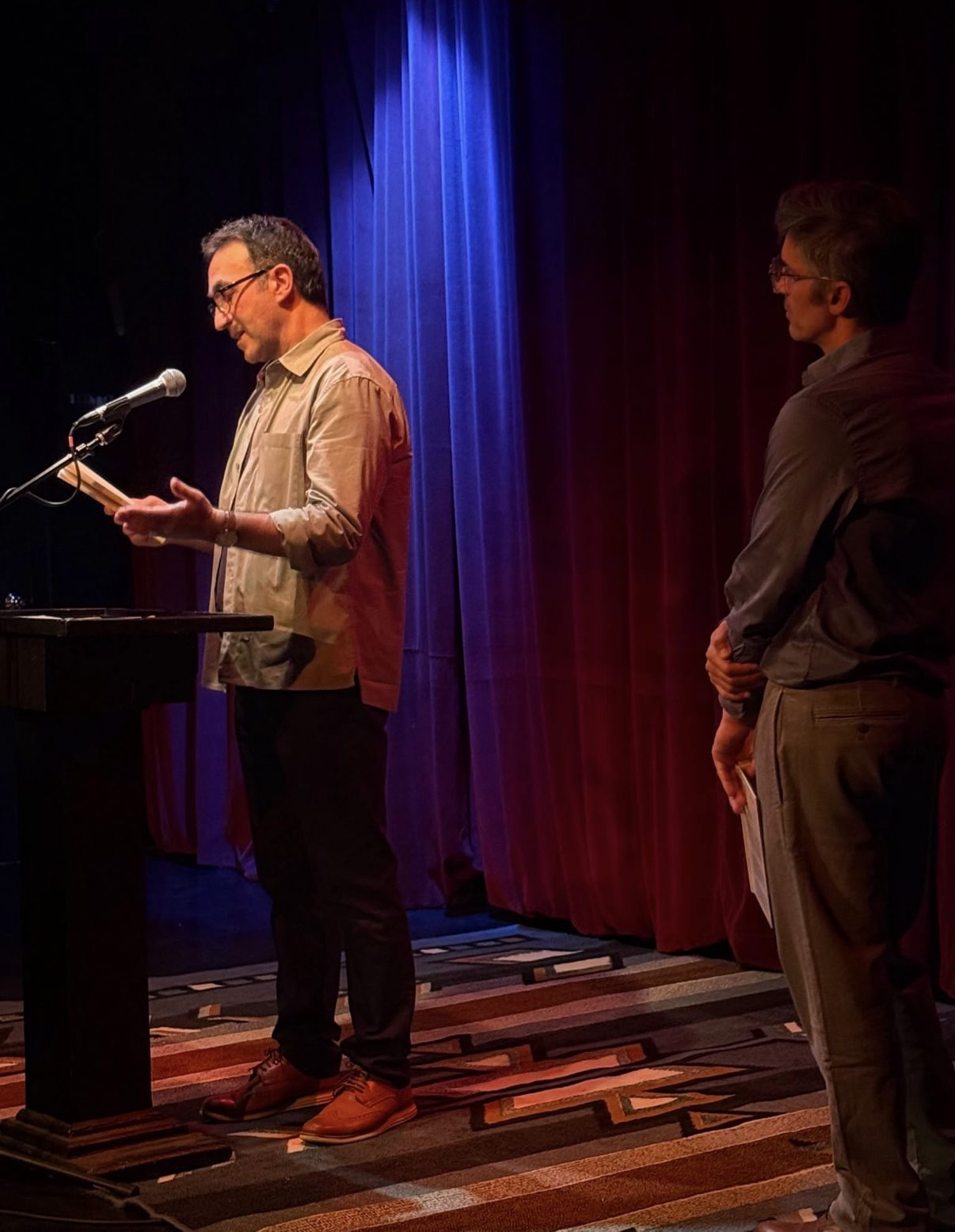 A fellow reader at the podium as Blas Falconer looks on